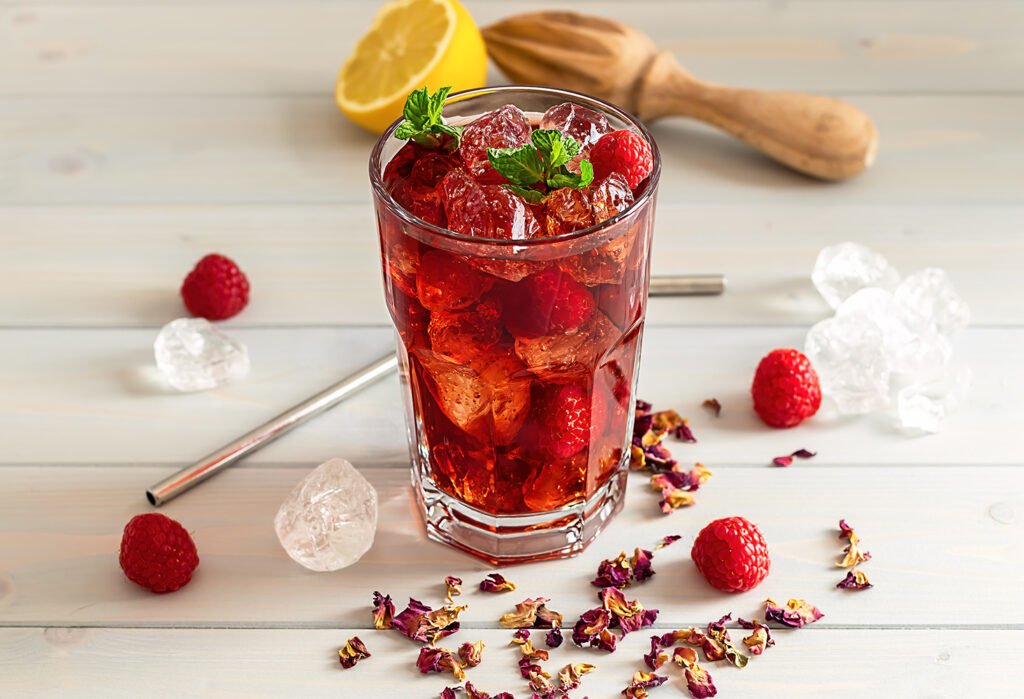 hibiscus ice tea or karkade lemonade with raspberries, mint, and lemon on a wooden table.